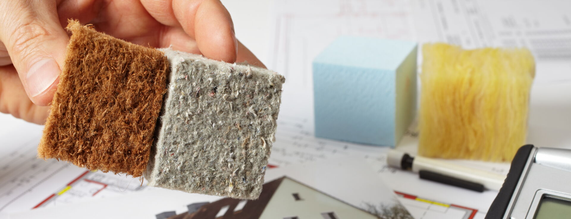 INTEGA Meinersdorf - Ingenieurbüro für Planungstätigkeit der Bauüberwachung, Abnahme & Übergabe der Anlagen Hand hält Dämmstoffmuster über Thermografiebild eines Hauses, Taschenrechner und Pläne im Hintergrund. Energieberatung in Südsachsen.